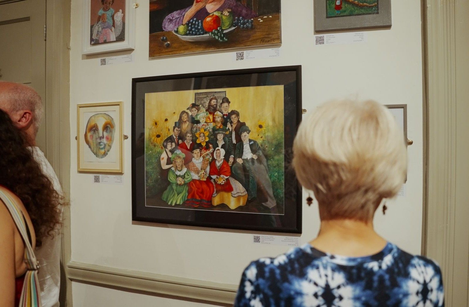 The back of a woman with short light coloured hair and a blue floral top who is looking at an artwork of a group of people..jpg The back of a woman with short light coloured hair and a blue floral top who is looking at an artwork of a group of people..jpg