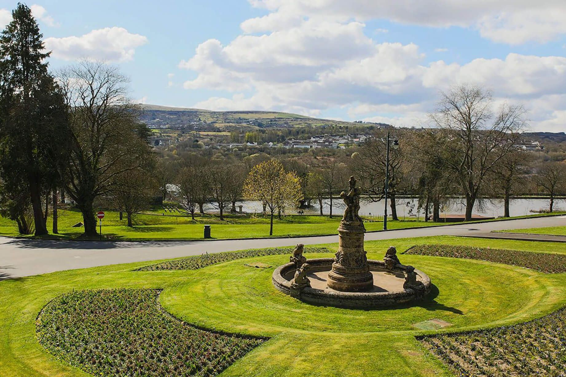 A circular stone fountain with no water surrounded by grass and plants with a lake and hills in the background.jpg